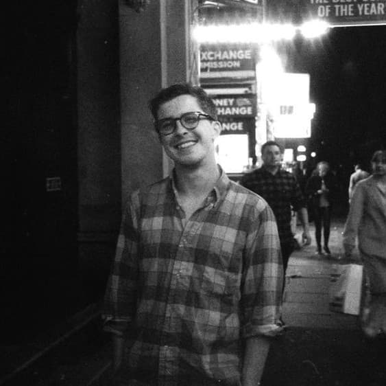 A smiling young man wearing glasses and a plaid shirt stands in front of a storefront with signs advertising currency exchange services.